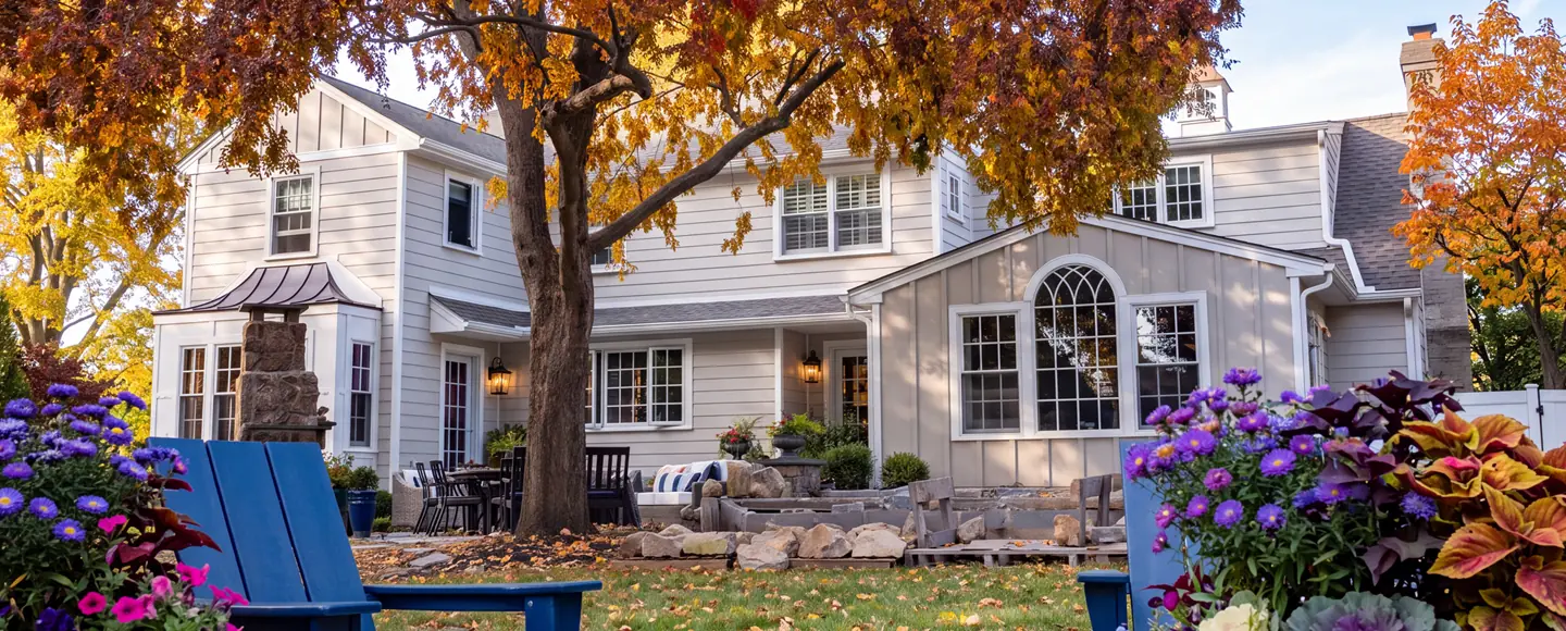 Exterior of a home featuring multiple windows and doors, viewed from a backyard patio surrounded by vibrant autumn trees, purple flowers, and blue Adirondack chairs.