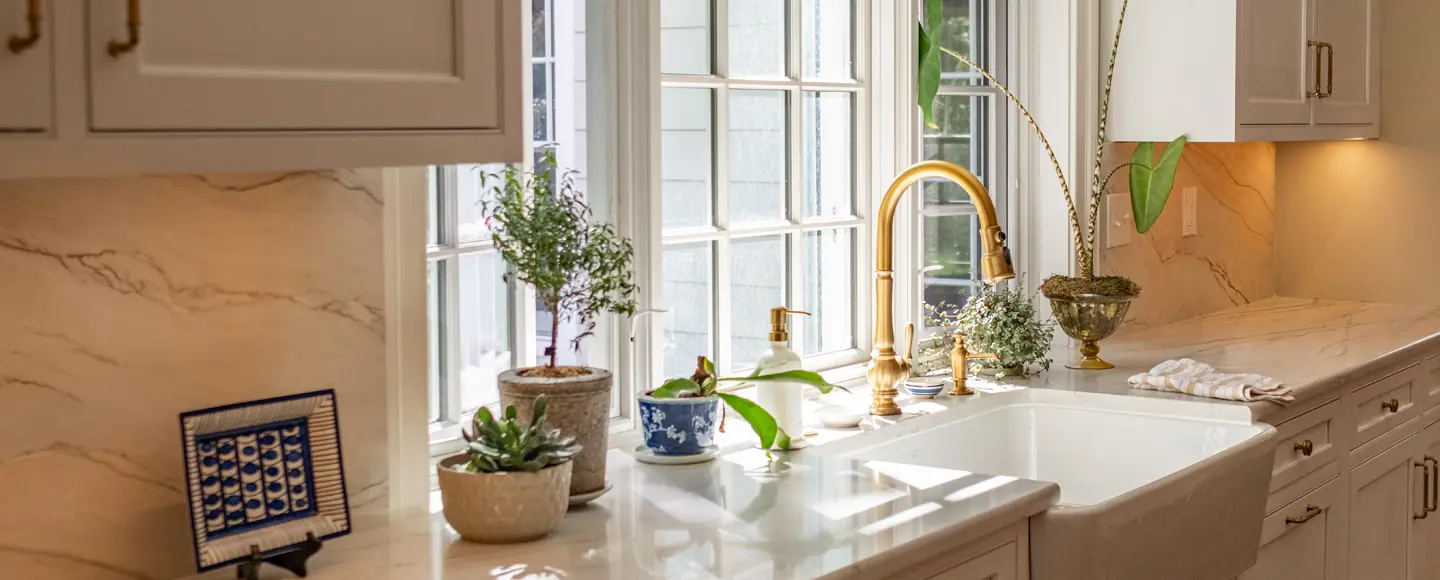 A bright kitchen interior showing a marble countertop, a white farmhouse sink, and a gold gooseneck faucet, with potted plants sitting in front of a large, sunlit multi-pane window.