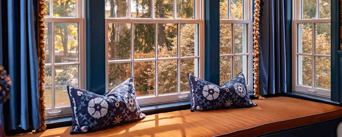A cozy indoor window seat with a rust-orange cushion and blue floral pillows, framed by large multi-pane windows looking out at autumn foliage.