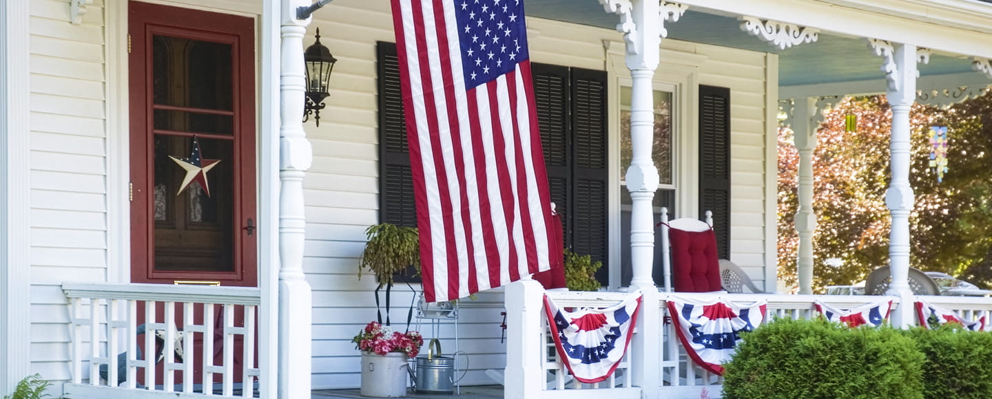 n American flag hangs from a white columned front porch decorated with patriotic bunting, featuring a prominent red storm door with a screen panel, black shutters, and rocking chairs.