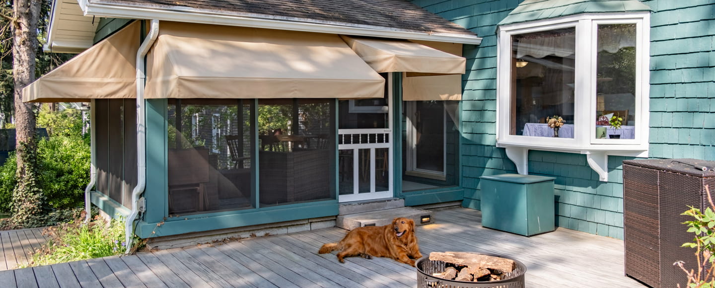 A sunny wooden deck featuring a screened-in porch with tan awnings, a golden retriever relaxing by a fire pit, and a large white bay window on a teal-shingled house.