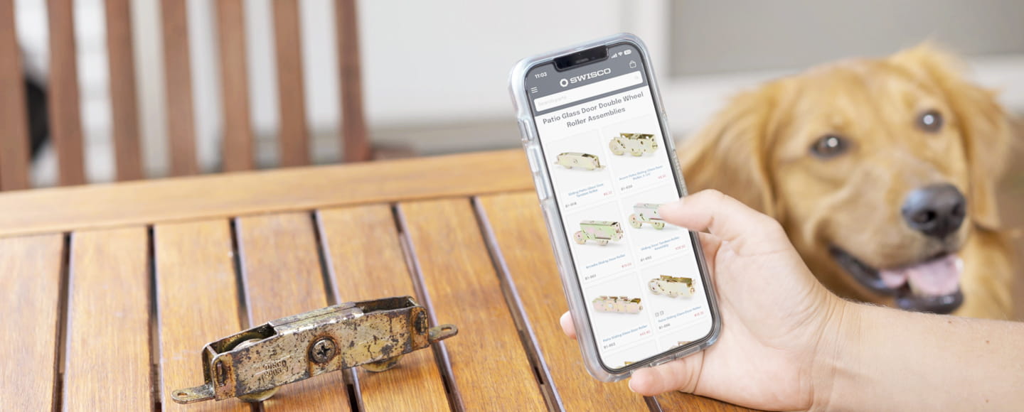 Hand holding an iPhone displaying patio glass door roller options on SWISCO.com next to an old, rusted metal roller assembly on a wooden table.