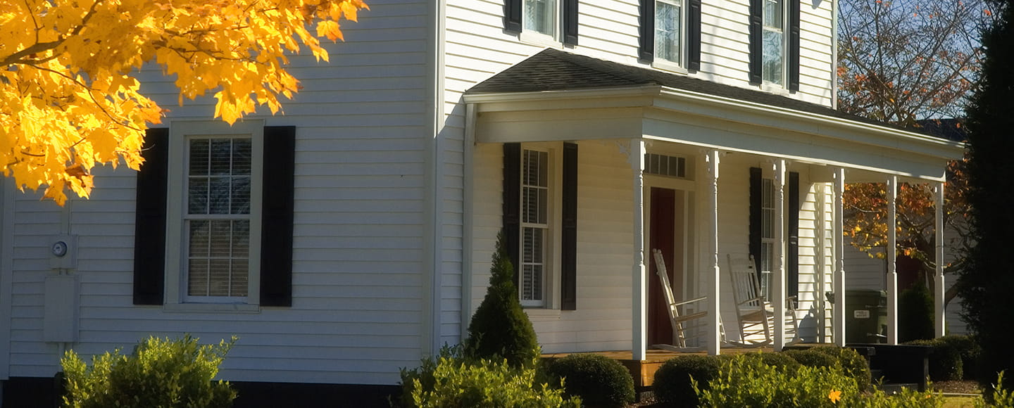 Exterior view of a white siding home featuring double-hung windows with black shutters, a front porch with rocking chairs, and bright yellow fall foliage.
