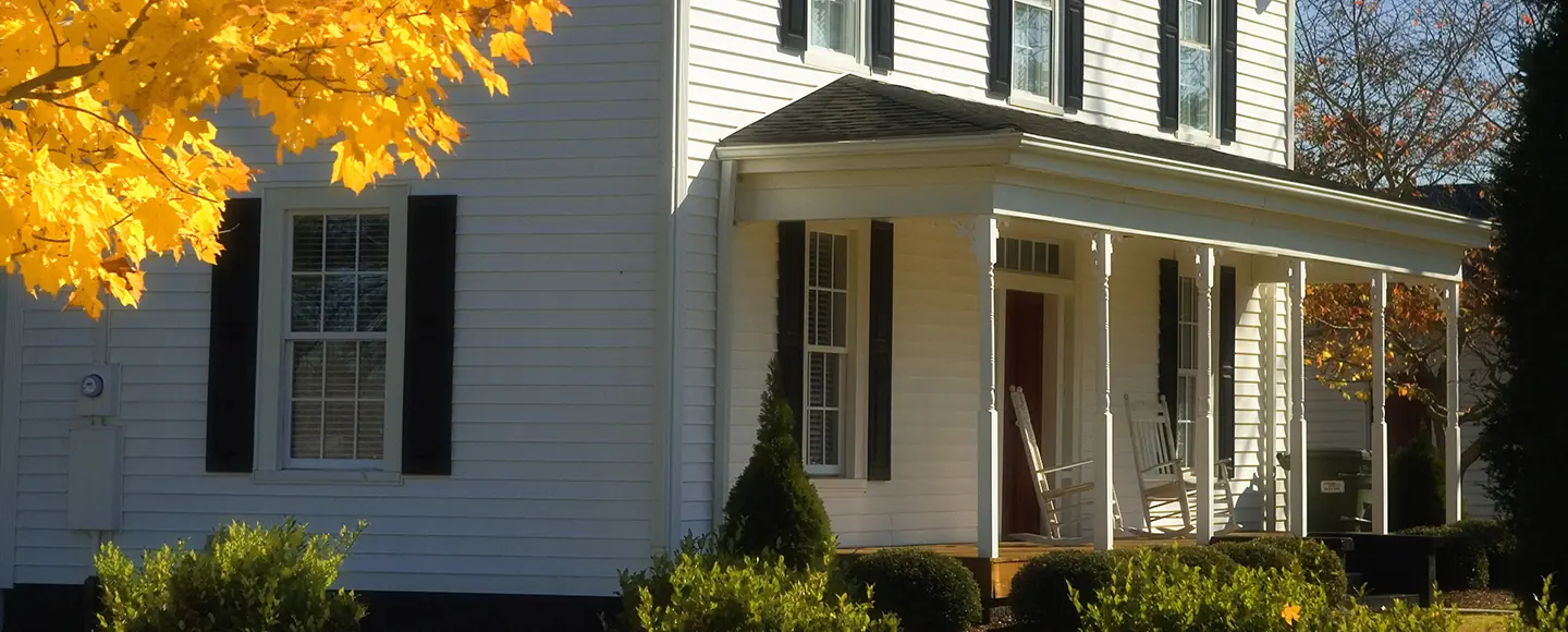 Exterior view of a white siding home featuring double-hung windows with black shutters, a front porch with rocking chairs, and bright yellow fall foliage.