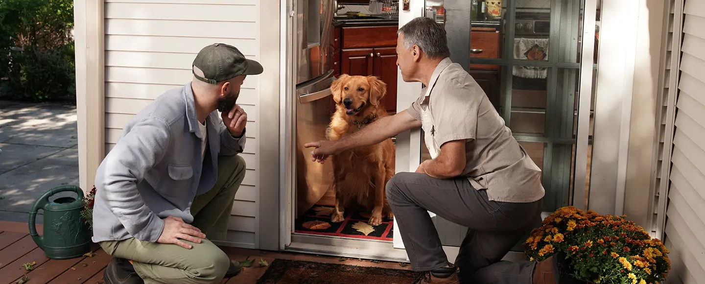 Man pointing to the bottom track of an open sliding screen door while explaining the repair to another man on an outdoor deck with a golden retriever watching.